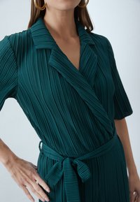 Smaragdgroene geplooide jurk met een kraag en halve mouwen. Heeft een strikceintuur in de taille en verticale textuur. Close-up van de bovenlichaam.