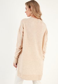 Pull en beige à manches longues avec des poignets et un ourlet côtelés. Texture douce, fentes latérales en bas et coupe décontractée avec un col arrondi.