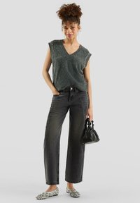 Woman with curly hair wears gray sleeveless knit top, black wide-leg jeans, embellished flats, and holds a small black handbag, standing casually.