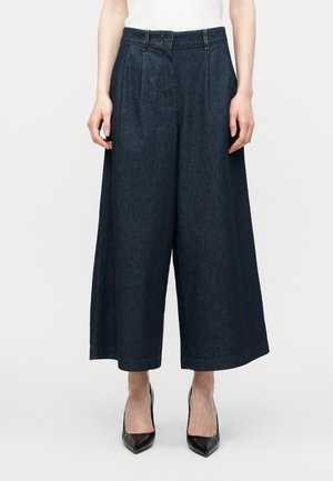 Pantalon large en denim foncé coupé court porté avec des talons noirs à bout pointu et un haut blanc sans manches, visible de la taille aux pieds.