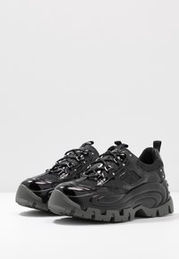 Černé sportovní boty s lesklým lakovaným povrchem, síťovými panely a silnými šedými gumovými podrážkami s hlubokým vzorkem pro lepší trakci.