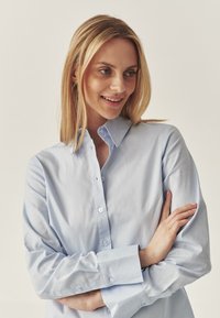 Camicia azzurra chiara con colletto strutturato, maniche lunghe e una consistenza liscia. Presenta bottoni bianchi e polsini leggermente arrotolati.