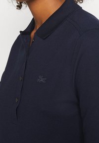 Navy blue polo shirt made of textured fabric with a buttoned placket and a ribbed collar, featuring a dark embroidered logo on the chest.
