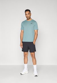 Camisa de manga corta de color verde teal claro con textura y cuello redondo, combinada con pantalones cortos oscuros. El modelo lleva zapatillas blancas y calcetines, posando sobre un fondo simple.