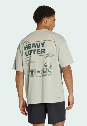 Muž v béžovém tričku "Heavy Lifter" s fitness motivy a ikonami na zádech, v černých šortkách, boční profil na jednoduchém pozadí.