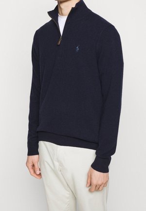 Homme portant un pull bleu marine à fermeture éclair quart de zip avec un petit logo sur la poitrine et un pantalon blanc, debout devant un fond clair uni.