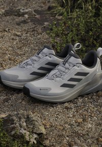 Lichtgrijze sportschoenen met zwarte accenten, een gestructureerde bovenkant en een flexibele zool, voorzien van een GORE-TEX-label voor waterbestendigheid.
