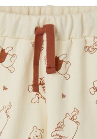 Cremefarbene Sweatpants mit einem braunen Kordelzug, versehen mit einem Allover-Druck von Cartoonbären und Bienen. Weicher, strukturierter Stoff.