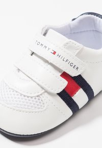 Białe skórzane sneakersy z siateczkowymi panelami, rzepem, czerwono-granatowymi paskami oraz logo "TOMMY HILFIGER" na rzepie.