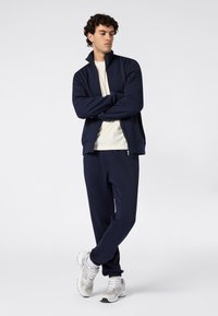Jonge man die met gekruiste armen staat, gekleed in een marineblauwe zip-up jas, bijpassende trainingsbroek, witte sneakers en een beige T-shirt.