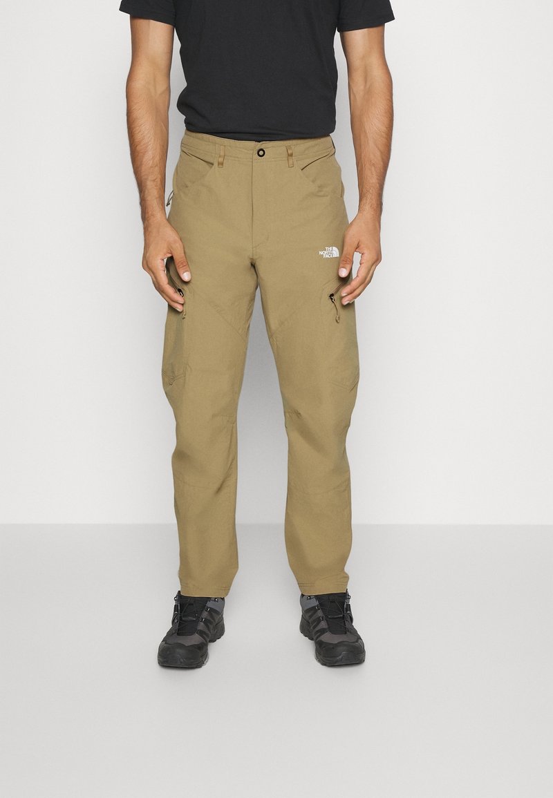 Homme portant un pantalon d’extérieur beige clair avec poches zippées et des chaussures de randonnée noires, debout devant un fond uni et clair.