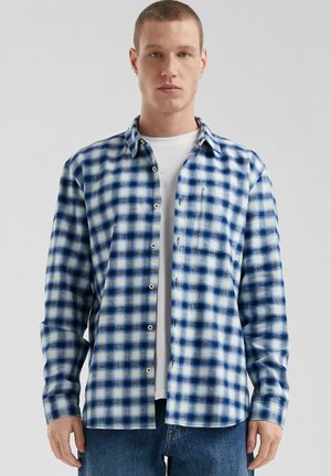 Blauw-wit geruite flanellen shirt met lange mouwen, knoopsluiting aan de voorkant en een linkse borstzak, gedragen over een wit T-shirt. Casual ontwerp.