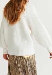 Witte gebreide trui met een gestructureerd patroon en ballonmouwen, gecombineerd met een gouden, bloemrijke plissé rok. Zacht, warm materiaal zichtbaar.