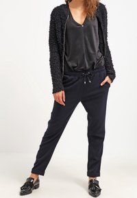 Svart, fluffig cardigan över en mörk, silkeslen topp; kombinerad med marinblå joggerbyxor och svarta loafers; avslappnad passform, casual design.