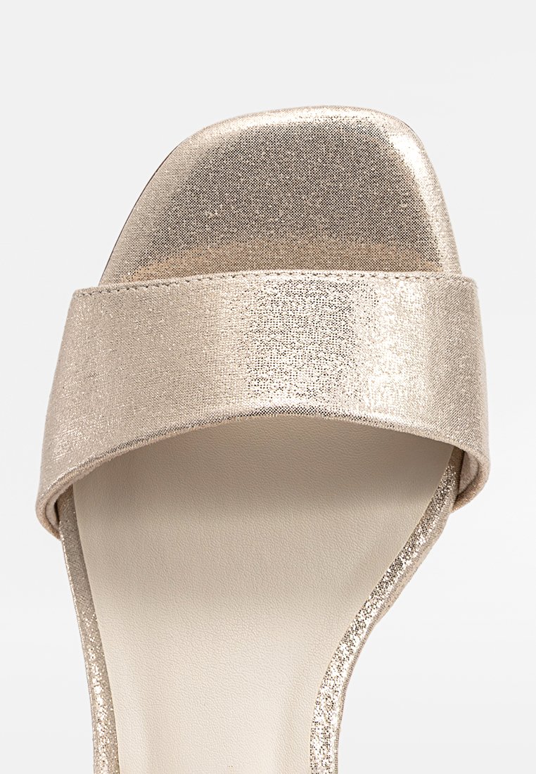 Vista frontale di un sandalo aperto d'oro metallico con una larga cintura su una superficie chiara.