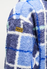 Υφασμα με μπλε και μοβ καρό σχέδιο, αφράτο, με λευκές λεπτομέρειες και καφέ δερμάτινο ταμπελάκι με την ένδειξη «ROXY» ραμμένο στην επιφάνεια.