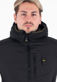 Giacca nera con cappuccio e colletto alto, chiusura con zip, dettagli giallo discreti e tasca sul petto con etichetta logo. Texture liscia.