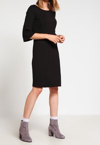 Czarna sukienka z rękawami o długości trzy czwarte, dopasowana do sylwetki, do kolan. Wykonana z miękkiego materiału i z okrągłym dekoltem. W zestawie z szarymi botkami za kostkę.