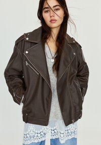 Junge Frau mit langen Haaren, die eine dunkelbraune Lederjacke über einem hellgrauen Pullover mit Spitzenbesatz und blauen Jeans trägt, mit den Händen in den Taschen.