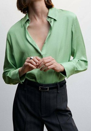 Femme portant une chemise vert clair boutonnée rentrée dans un pantalon noir taille haute avec une ceinture noire, ajustant sa bague en argent.