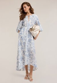 Floral maxi dress in light blue and white, featuring three-quarter sleeves, a tie neckline, and a high-low hem. Accompanied by a cream handbag.