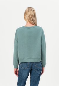 Hanorac verde deschis cu decolteu rotund și mâneci lungi. Are o tăietură relaxată și este asortat cu blugi de denim albaștri.