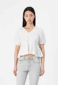 Fehér, rövid ujjú, peplumos felső V-kivágással. Ráncolt anyaggal és enyhén rövidített fazonnal rendelkezik, világosszürke farmernadrággal viselve.