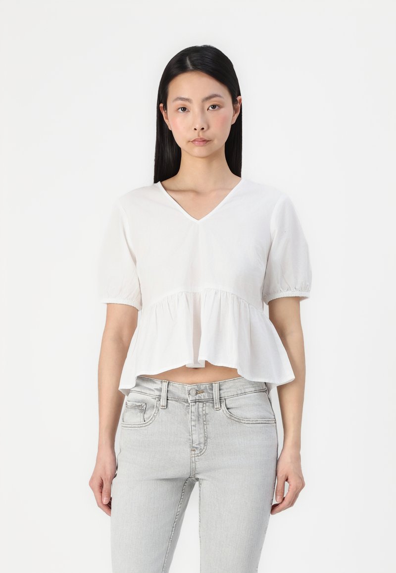 Fehér, rövid ujjú, peplumos felső V-kivágással. Ráncolt anyaggal és enyhén rövidített fazonnal rendelkezik, világosszürke farmernadrággal viselve.