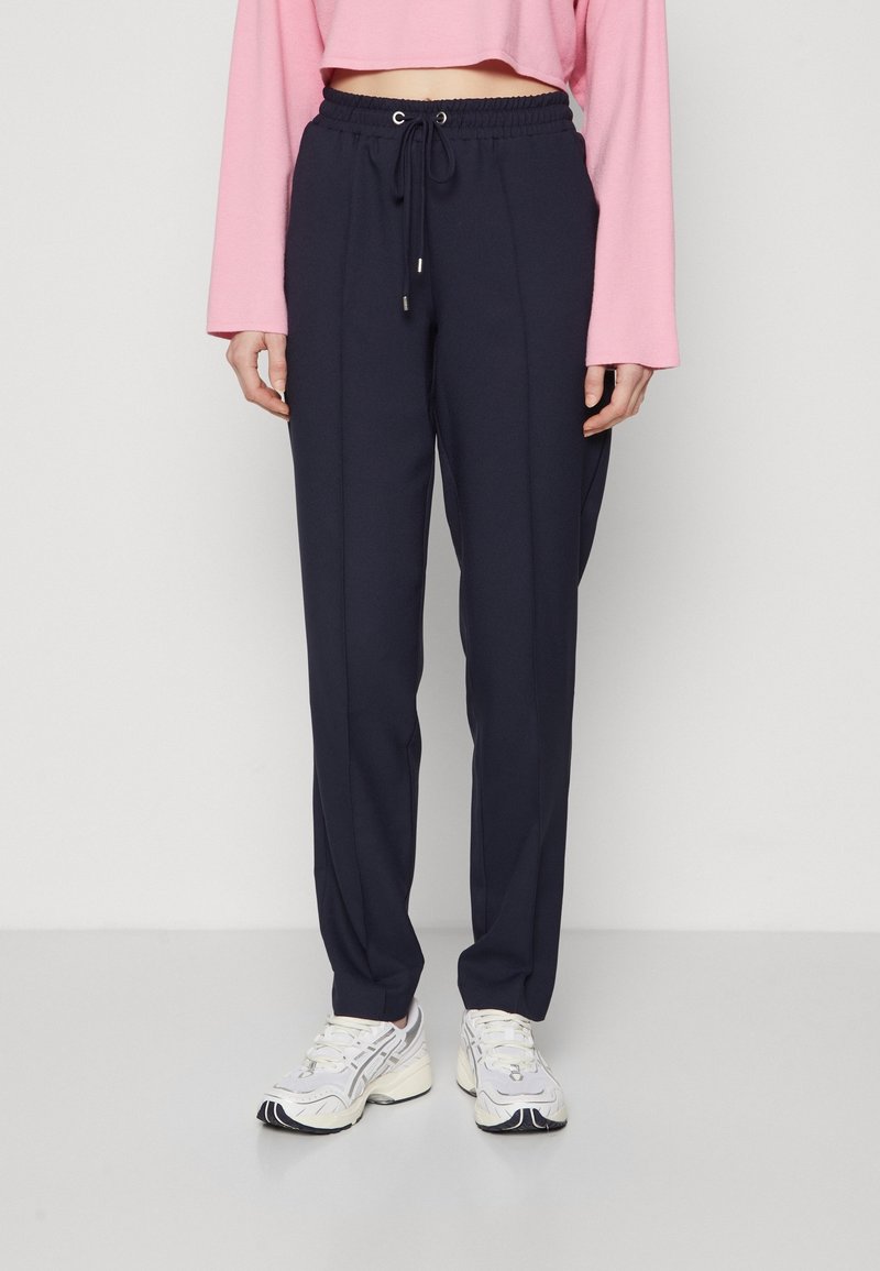 Personne portant un pantalon à cordon bleu marine, des baskets blanches et un crop top rose à manches longues, debout sur un fond clair uni.