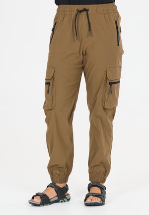 Bruine cargobroek met elastische tailleband en pijpboorden, meerdere zakken met rits, gedragen met zwarte outdoorsandalen op een witte achtergrond.