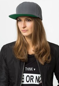 Casquette snapback grise avec une visière plate verte, texture de tissu doux et couture détaillée. Portée avec une veste bomber noire et un t-shirt.