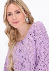 Cardigan tricotat în nuanță de liliac, cu un model texturat din dantelă, având cinci butoane mari de culoare purpurie pe față și accente subtile de sclipici.