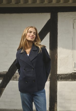 Marineblauwe dubbelzijdige jasje met grote zwarte knopen, gemaakt van wol. Het heeft een klassieke kraag en is gecombineerd met blauwe jeans.