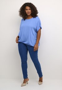 Světle modré volné tričko s krátkým rukávem skombinované s tmavě modrými skinny džíny. Model má na sobě béžové kožené muly na podpatku. Čisté pozadí.