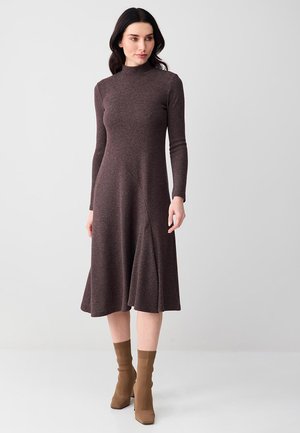 Femme portant une robe midi marron foncé à manches longues et col montant, associée à des bottines marron clair, posant devant un fond gris uni.