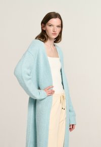 Light blue long cardigan made of soft, fluffy knit material with an open front, wide sleeves, and ribbed texture. Paired with a white tank top.