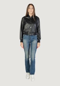 Giacca bomber nera in pelle con design accorciato, chiusura a bottone a pressione, polsini a coste, abbinata a jeans azzurri e sneakers bianche.