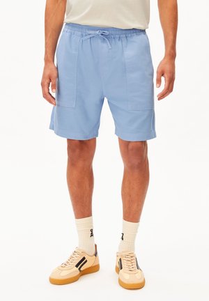 Homme portant un short décontracté bleu clair avec des poches avant, des chaussettes crème et des baskets beige avec des rayures noires, debout devant un fond blanc.