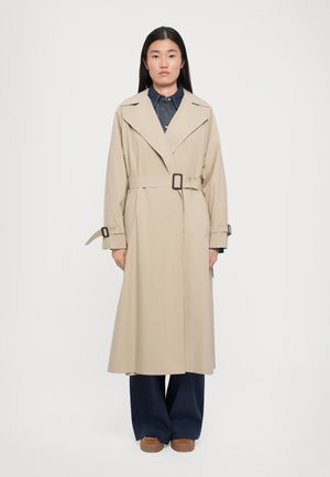 Trench coat beige con vita a cintura, ampi revers e maniche lunghe. Indossato sopra una camicia di jeans, abbinato a pantaloni blu navy e scarpe marroni.