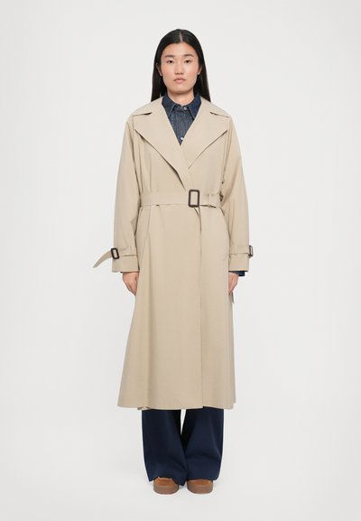 Beige trenchcoat med bælte i taljen, brede klapper og lange ærmer. Båret over en denimskjorte, parret med marineblå bukser og brune sko.