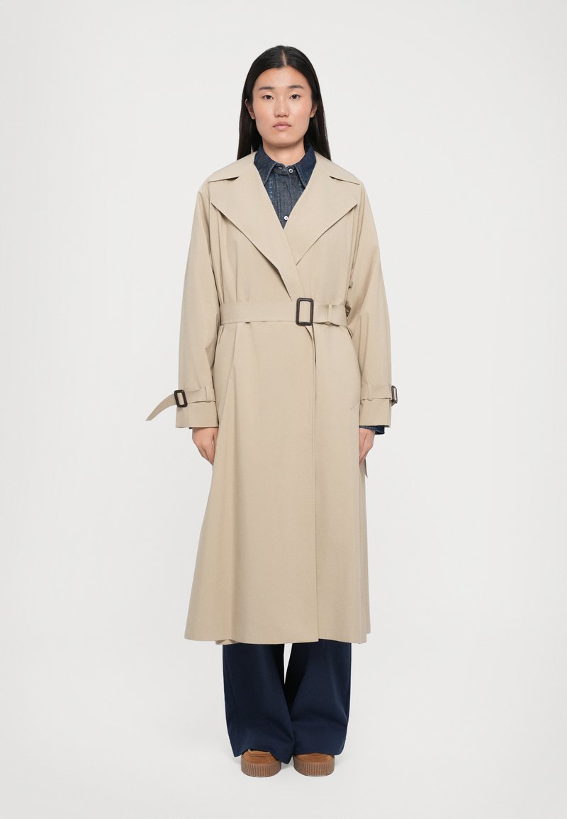 Beige trence coats ar jostu jostu, platām atlociem un garām piedurknēm. Uzvilkta virs džinsa krekla, saskaņota ar tumši ziliem biksēm un brūnām kurpēm.
