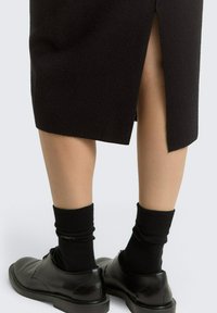Zwarte wollen rok met een split aan de achterkant, gecombineerd met zwarte sokken en glanzende zwarte schoenen. De textuur lijkt glad en gestructureerd.