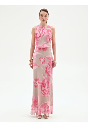 Donna con abito senza maniche, lungo fino al pavimento, con grande motivo floreale rosa su sfondo beige, in piedi davanti a uno sfondo bianco semplice.