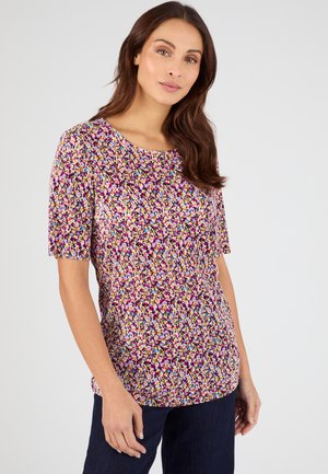 Mujer con una blusa de manga corta, con estampado floral multicolor y pantalones oscuros, de pie contra un fondo claro y liso.