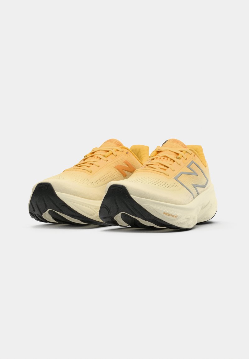 Un par de zapatillas de running New Balance amarillas y crema con suelas negras, posicionadas una al lado de la otra sobre un fondo blanco.