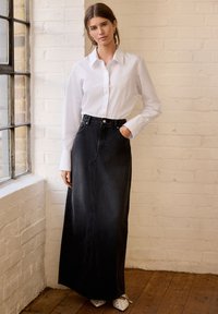 Svart denimmaxikjol med en framslits, hög midja och kontrasterande sömmar. Kombinerad med en vit knapp-up skjorta och spetsiga skor.
