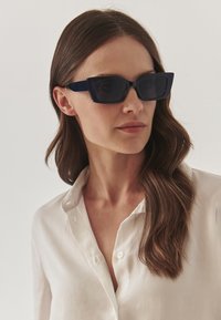 Tamsiai mėlyni stačiakampiai akiniai nuo saulės su tamsiais lęšiais ir storu rėmu, derinami su balta marškinėlių apykakle. Lygus paviršius, modernus dizainas.