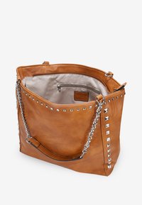 Bolso tote de cuero marrón claro con tachuelas piramidales plateadas a lo largo del borde superior; cuenta con un asa de cadena y un bolsillo interior con cremallera.