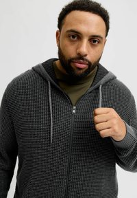 Haut de sweatshirt zippé gris avec un motif texturé, accents gris clair contrastants sur la capuche et la fermeture éclair, porté sur un t-shirt vert foncé.
