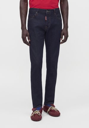 Homme portant un jean bleu foncé, une chemise rouge et des baskets rouges avec des lacets bleus et beiges, debout devant un fond uni.